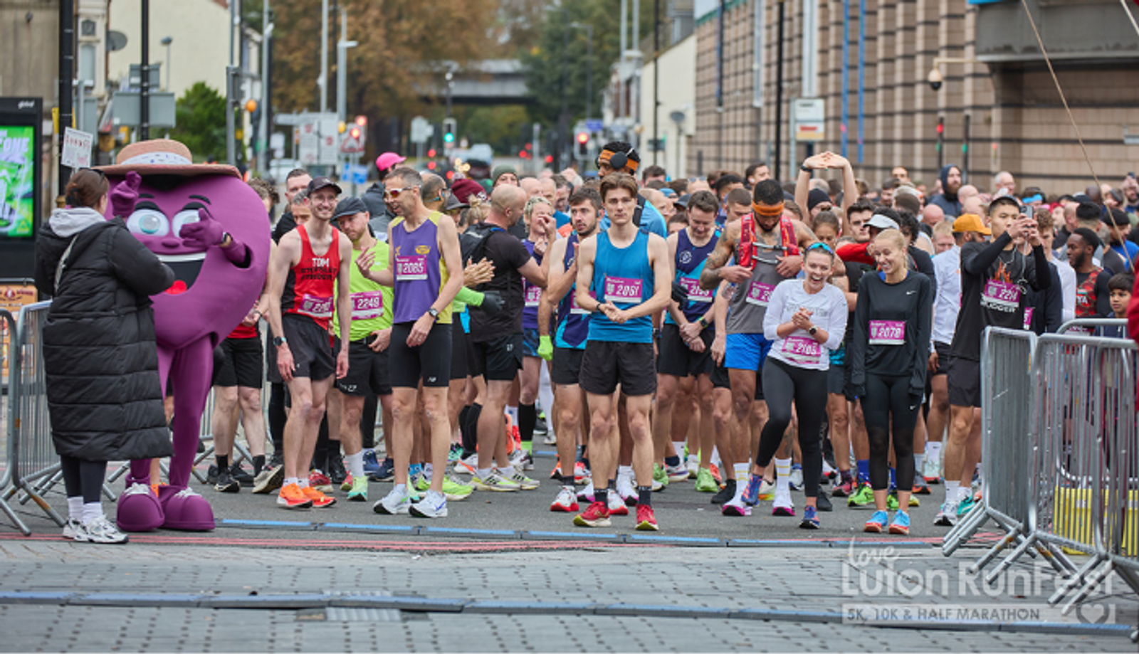 A large group of runners gathers at the starting line of a road race in a city. A person in a purple mascot costume stands to the left, while participants wait and stretch before beginning the event.