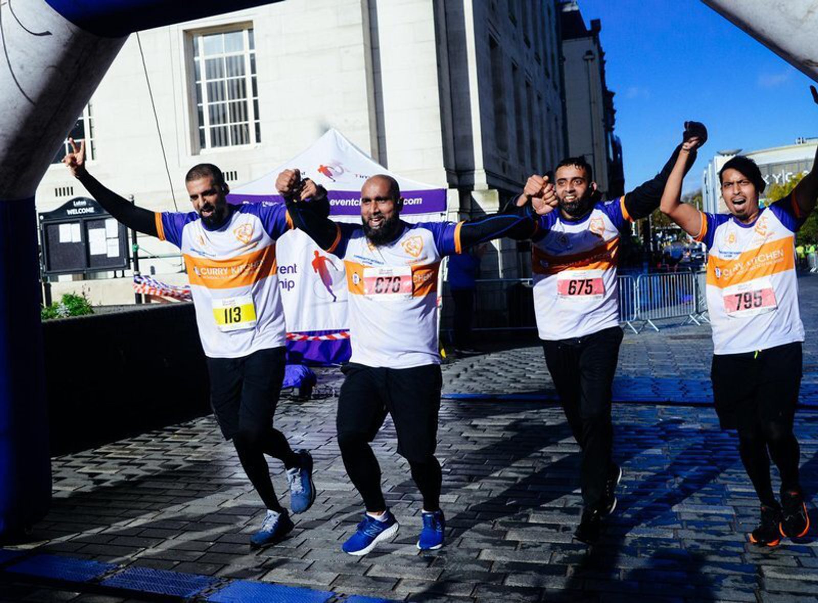 Four runners in athletic gear are holding hands and celebrating as they cross the finish line together during a race. The runners display joyful expressions, some have their arms raised in victory. They're wearing numbered bibs in a city street setting.