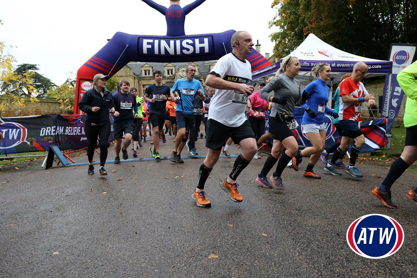 Runners cross the finish line at an outdoor race event, with an inflatable arch labeled "FINISH" overhead and trees and buildings visible in the background. The ground is wet, and people are dressed in athletic gear.