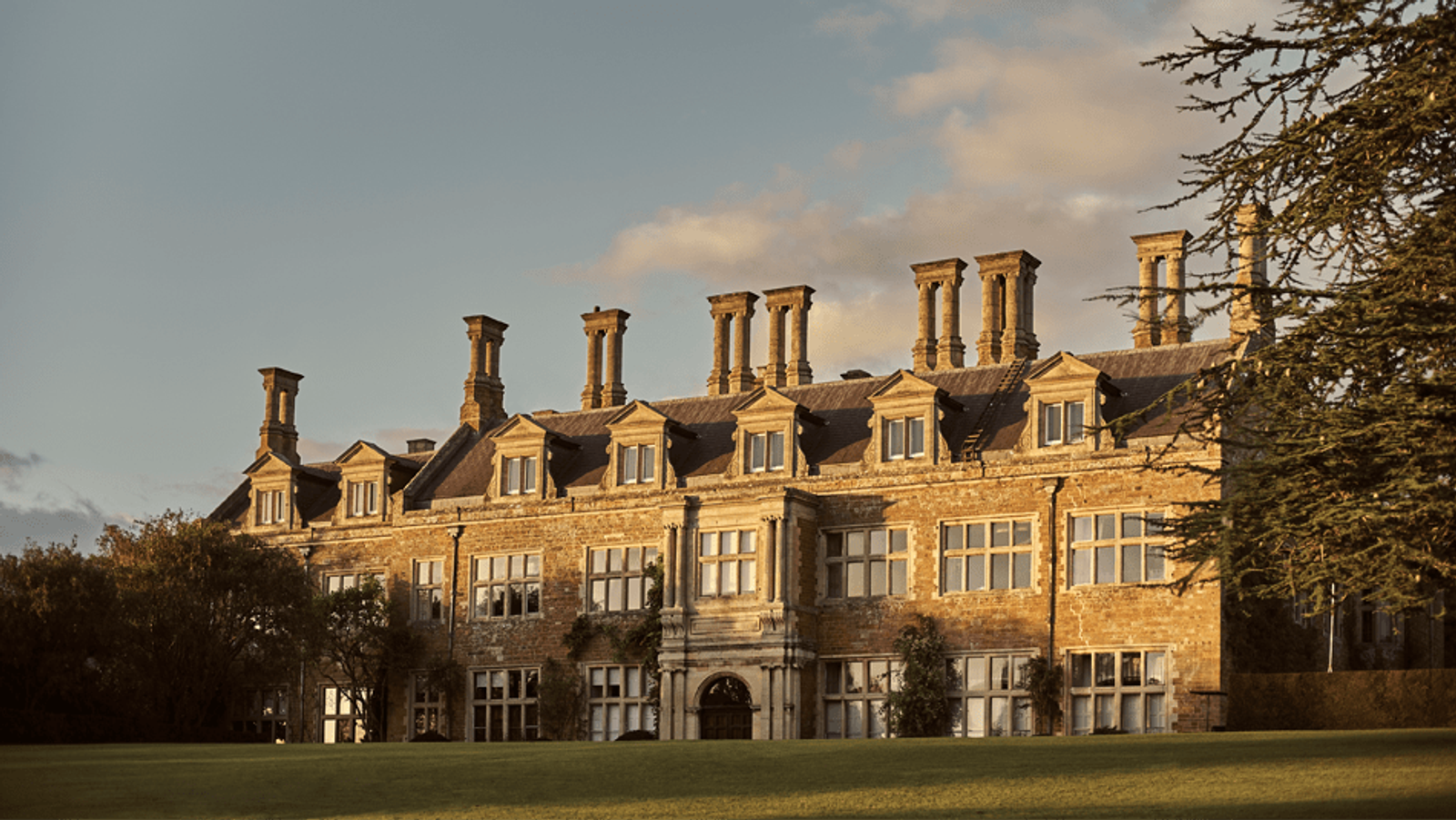 A historic, multi-story stone building with numerous chimneys and large windows, sits under a partly cloudy sky. The facade features ivy-covered walls and an arched entrance. A large tree and well-manicured green lawn surround the building.
