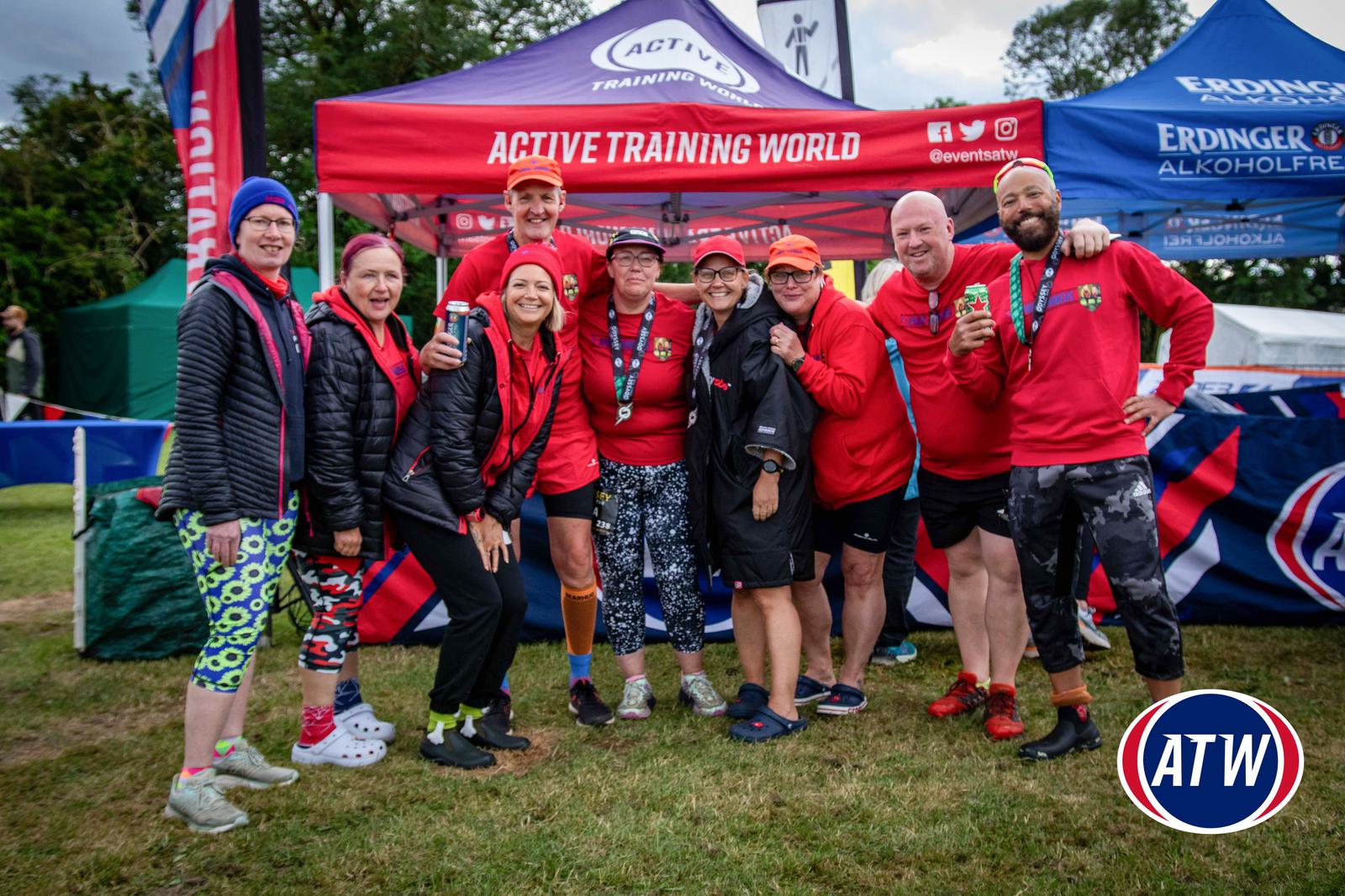 A group of people in red and black sportswear pose and smile in front of an "Active Training World" tent. They hold drinks and wear medals, indicating participation in an event. A logo with "ATW" is on the grass.