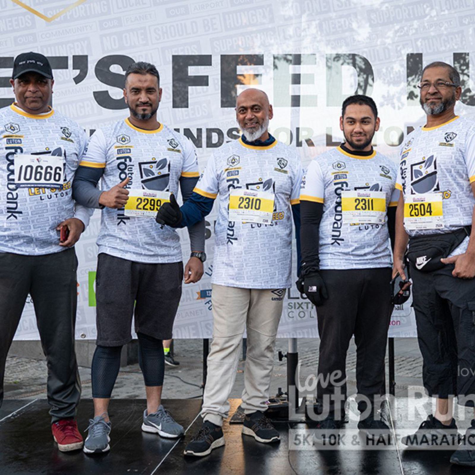 Five men wearing matching race shirts stand together in front of a banner at the Love Luton Run event. They each have race bibs with numbers and smile at the camera, showing a sense of camaraderie.