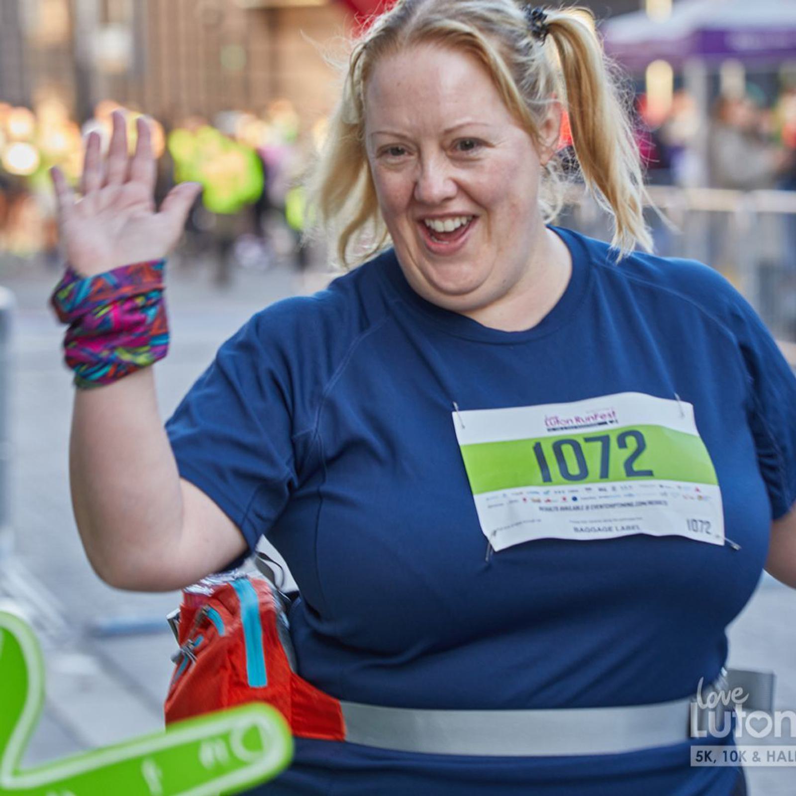 A woman with blonde hair in pigtails, wearing a blue running shirt with a race number, smiles and waves while participating in a race. The background shows other runners and event signage.