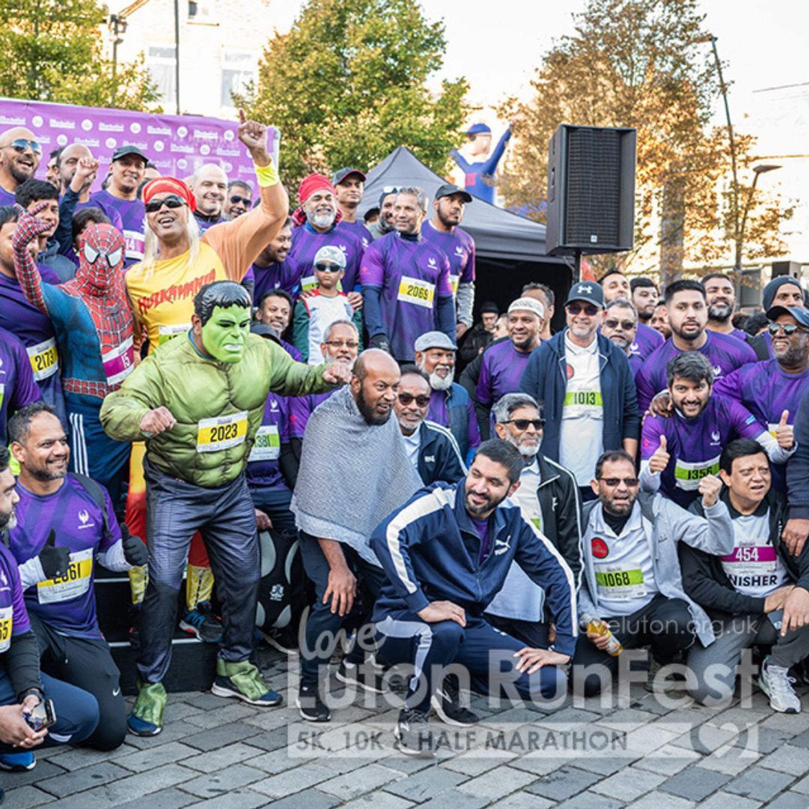 A group of runners dressed in purple shirts and various costumes, including superheroes, gather for a photo at the Love Luton RunFest. The setting is outdoors, with trees and buildings visible in the background.