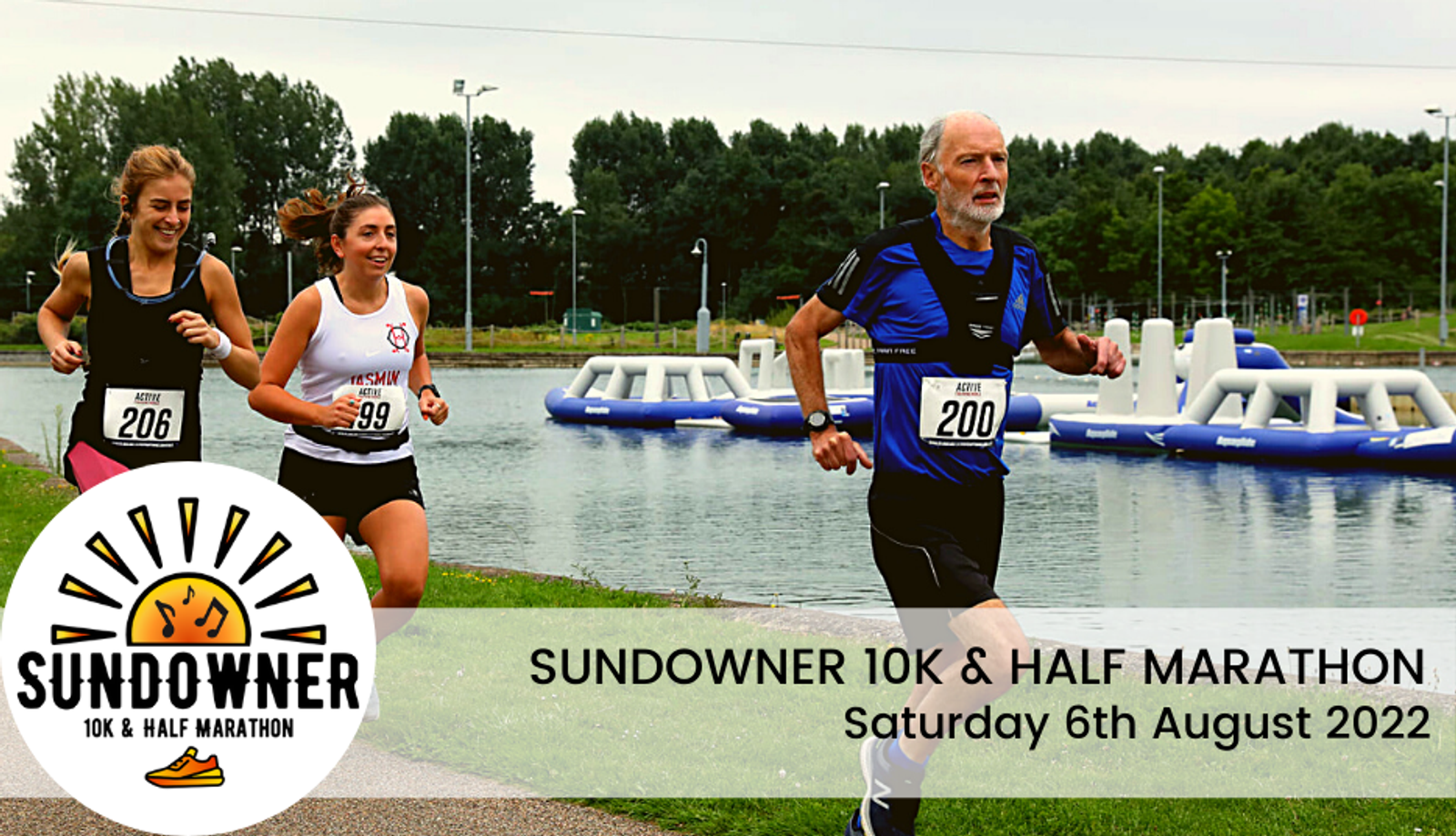 Three runners participate in the Sundowner 10K & Half Marathon beside a lake with inflatable obstacles. Two young women are in the foreground, wearing bib numbers 26 and 99, while an older man with bib number 200 runs slightly ahead. Event details overlay the image.