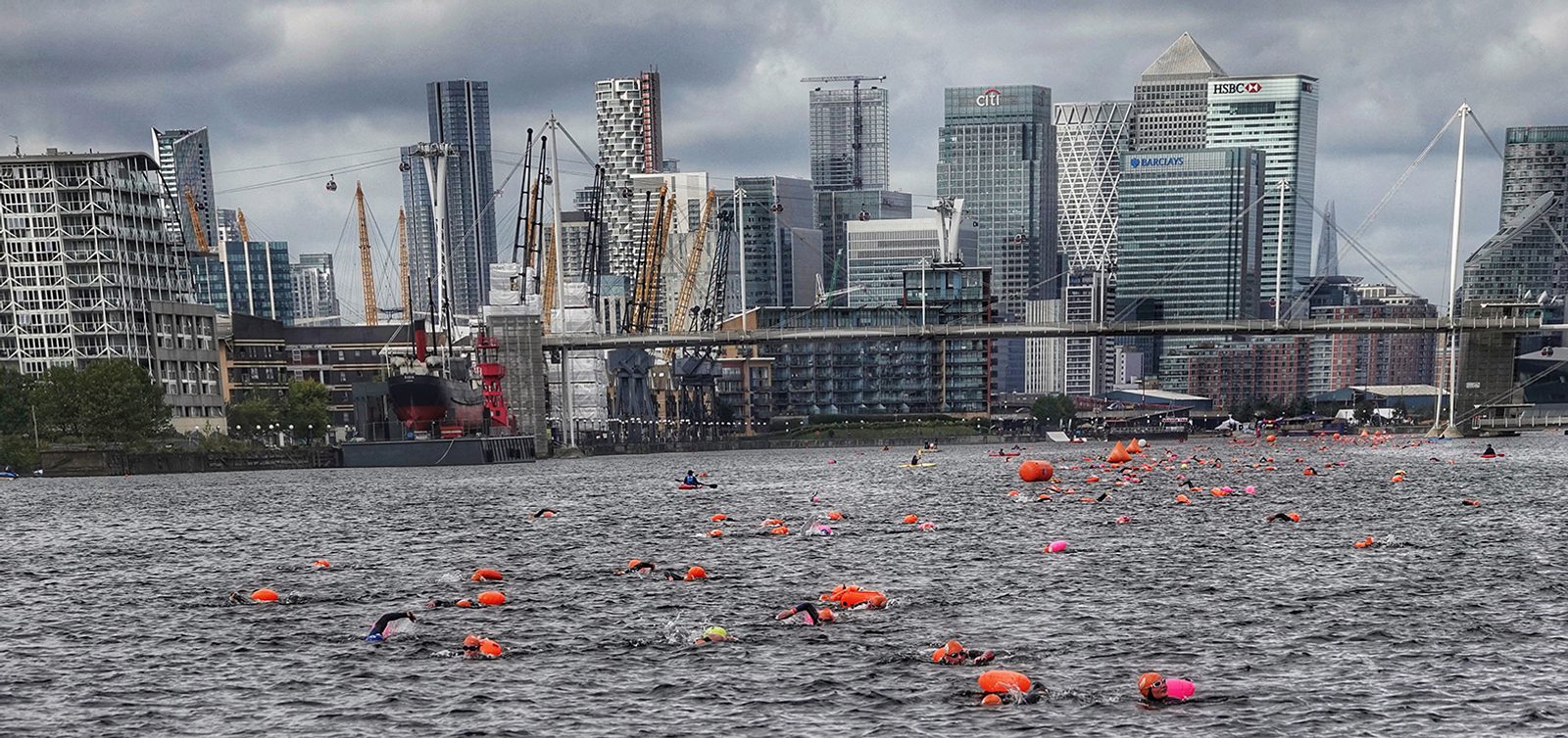 A large group of swimmers wearing orange swim caps and buoyancy aids participate in an outdoor event in a river or lake with tall skyscrapers and cranes in the background under a cloudy sky.