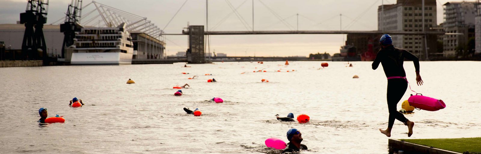 Swimmers in wetsuits and caps participate in an open water swimming event in a river with various brightly colored floating markers. A swimmer exits the water onto a grassy platform while others continue swimming. An industrial cityscape and bridge are in the background.