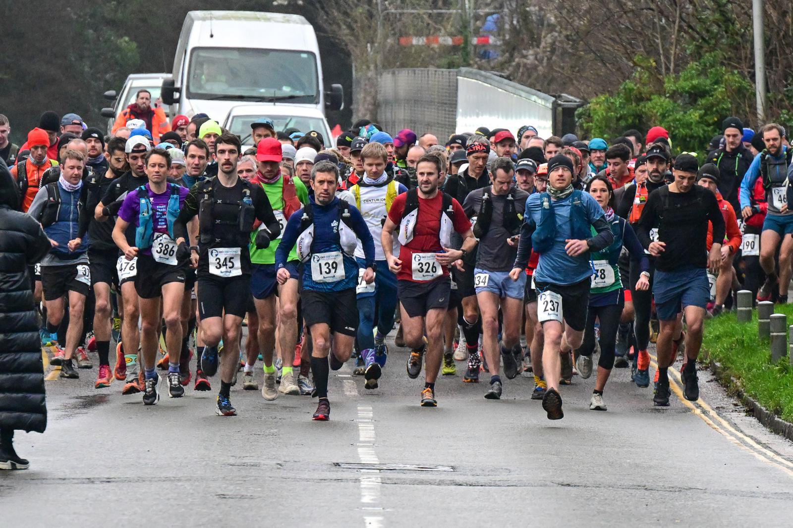 A large group of runners participating in a road race, wearing numbered bibs and athletic gear. They are focused, and a white truck is visible in the background. The weather appears cool and overcast, with some runners wearing long sleeves.