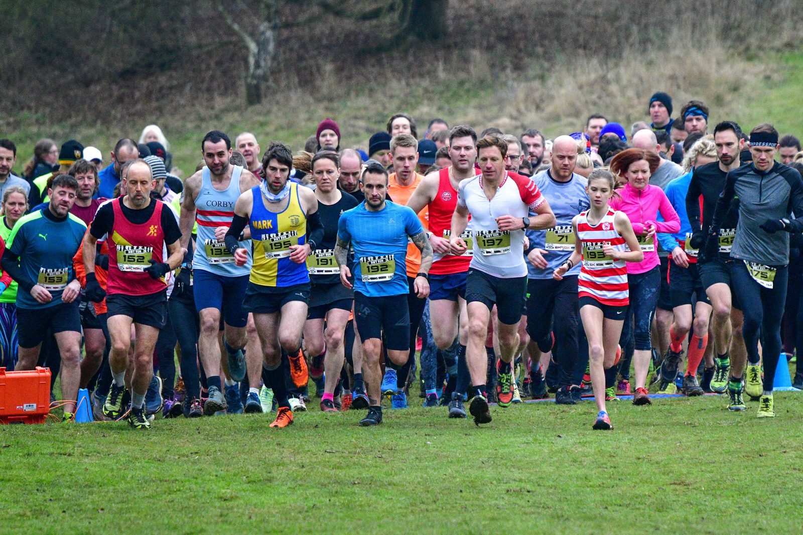 A large group of runners, wearing numbered bibs and diverse athletic attire, are starting a race on a grassy field. Spectators are visible in the background, dressed in winter clothing. The runners range in age and gender, all appearing focused and determined.