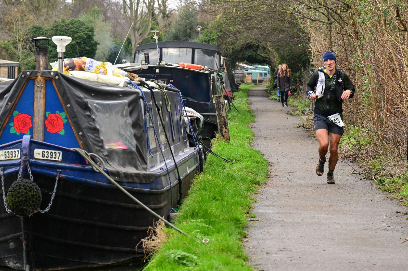 A runner in athletic gear jogs along a narrow path beside moored canal boats. The path is lined with bare trees, and a few people walk in the background. Overcast weather adds a moody atmosphere to the scene.