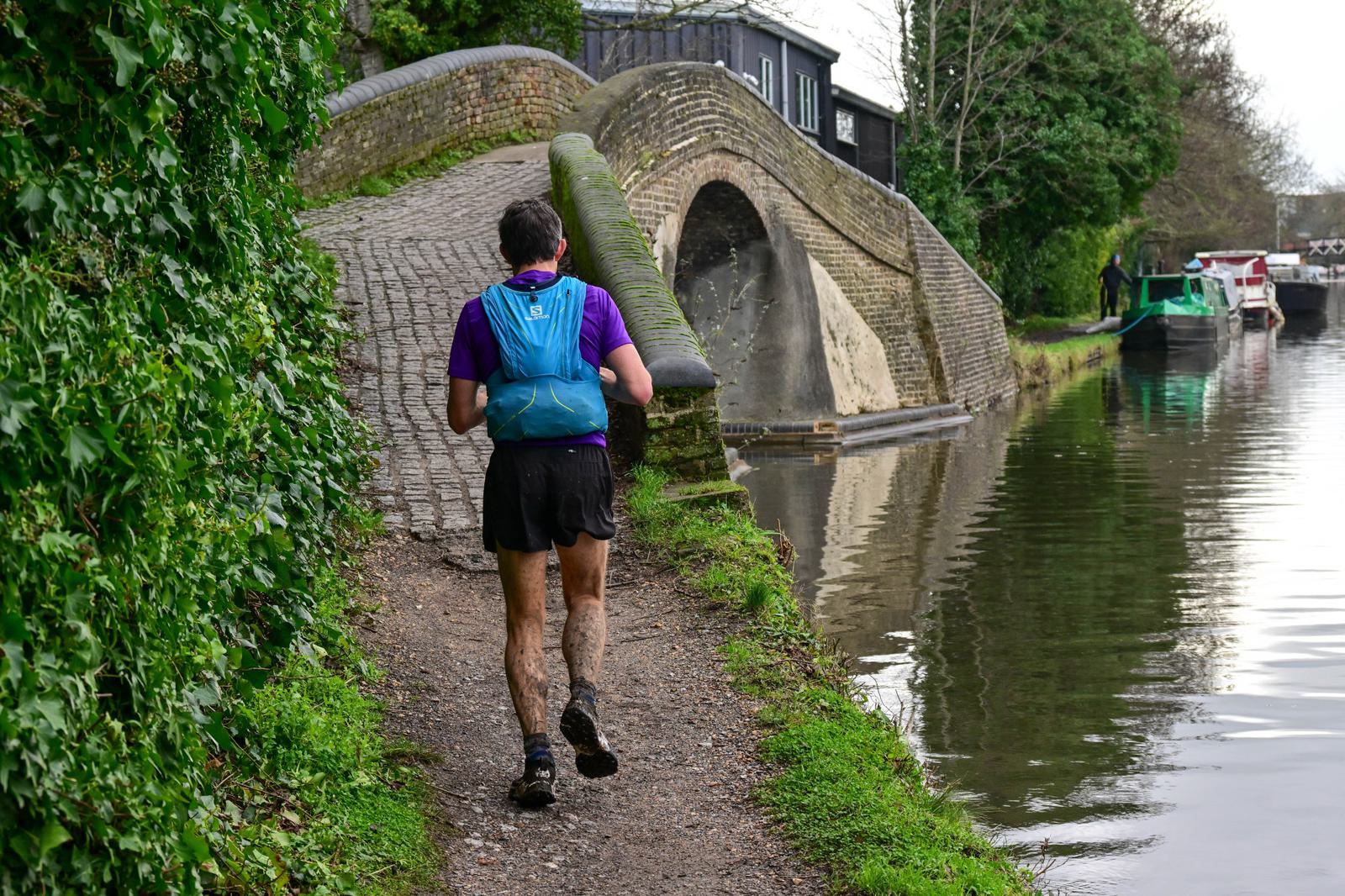 A person runs on a muddy path beside a canal, wearing a blue backpack and a purple shirt. A narrow, arching stone bridge is in the background, with lush greenery on the left and boats docked on the right.