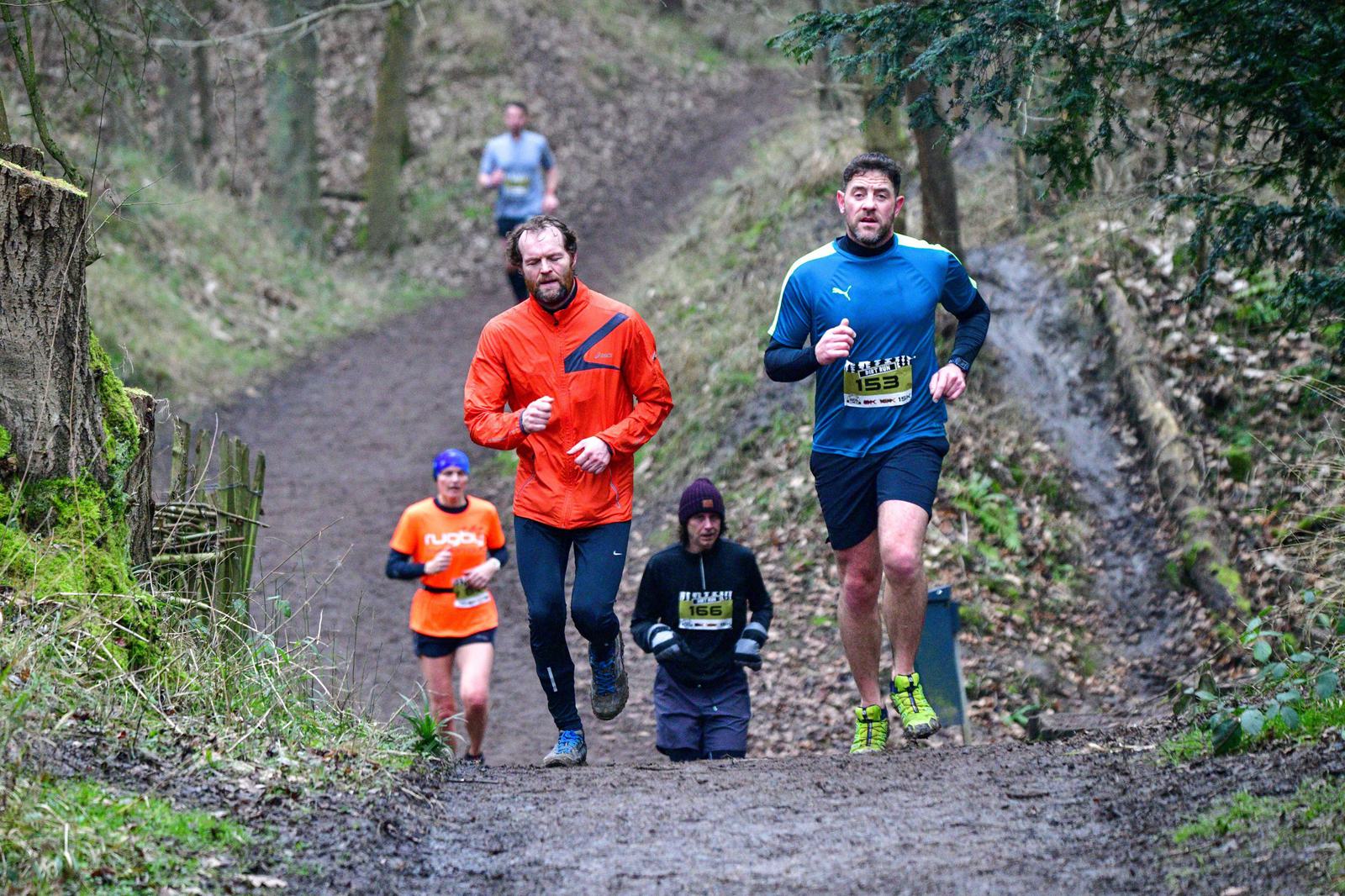 Four people are running uphill on a muddy trail through a wooded area. Two runners in front wear bright jackets—orange and blue. Two more runners are slightly behind, wearing darker attire. The scene conveys a sense of determination and camaraderie.