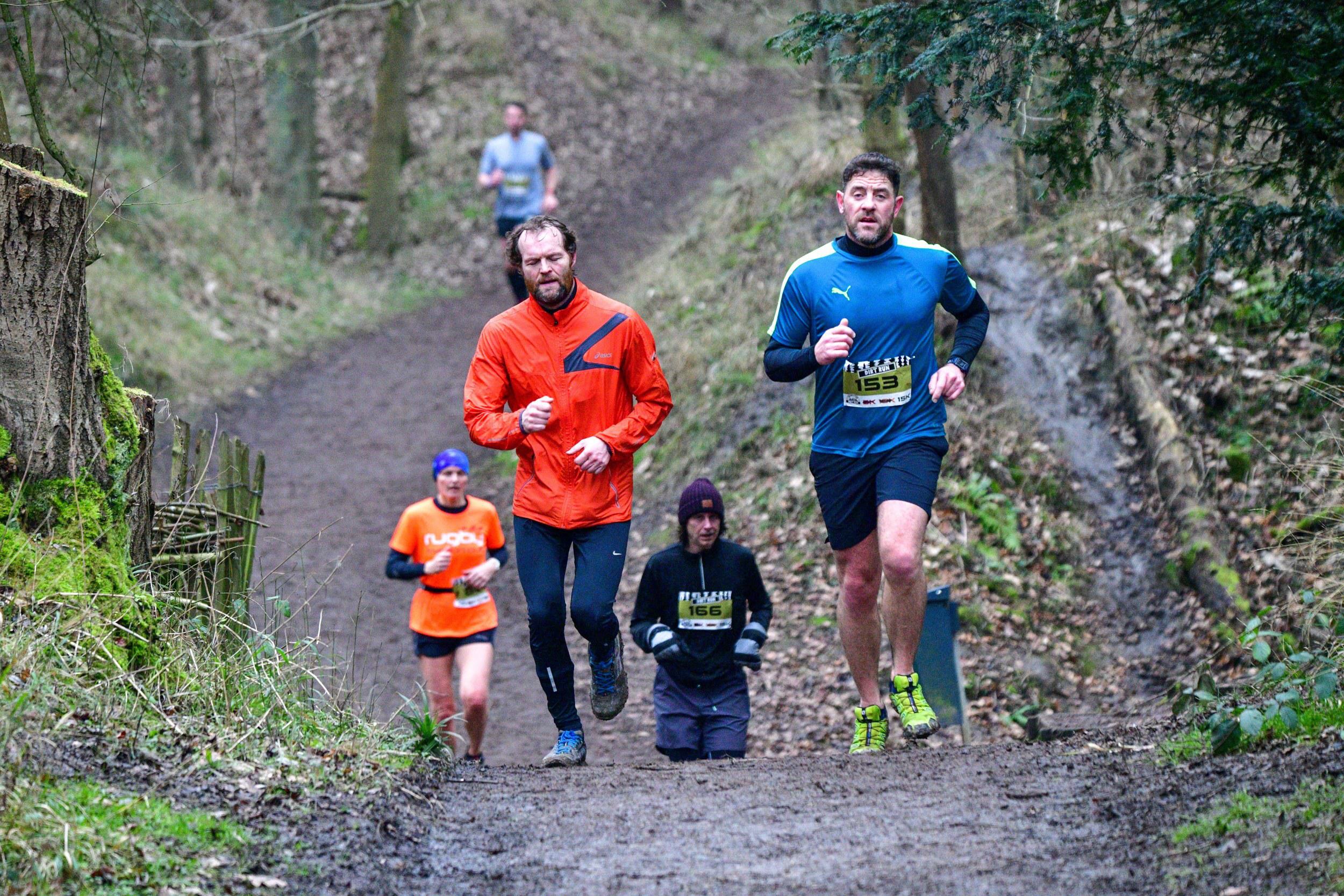 Four people are running uphill on a muddy trail through a wooded area. Two runners in front wear bright jackets—orange and blue. Two more runners are slightly behind, wearing darker attire. The scene conveys a sense of determination and camaraderie.