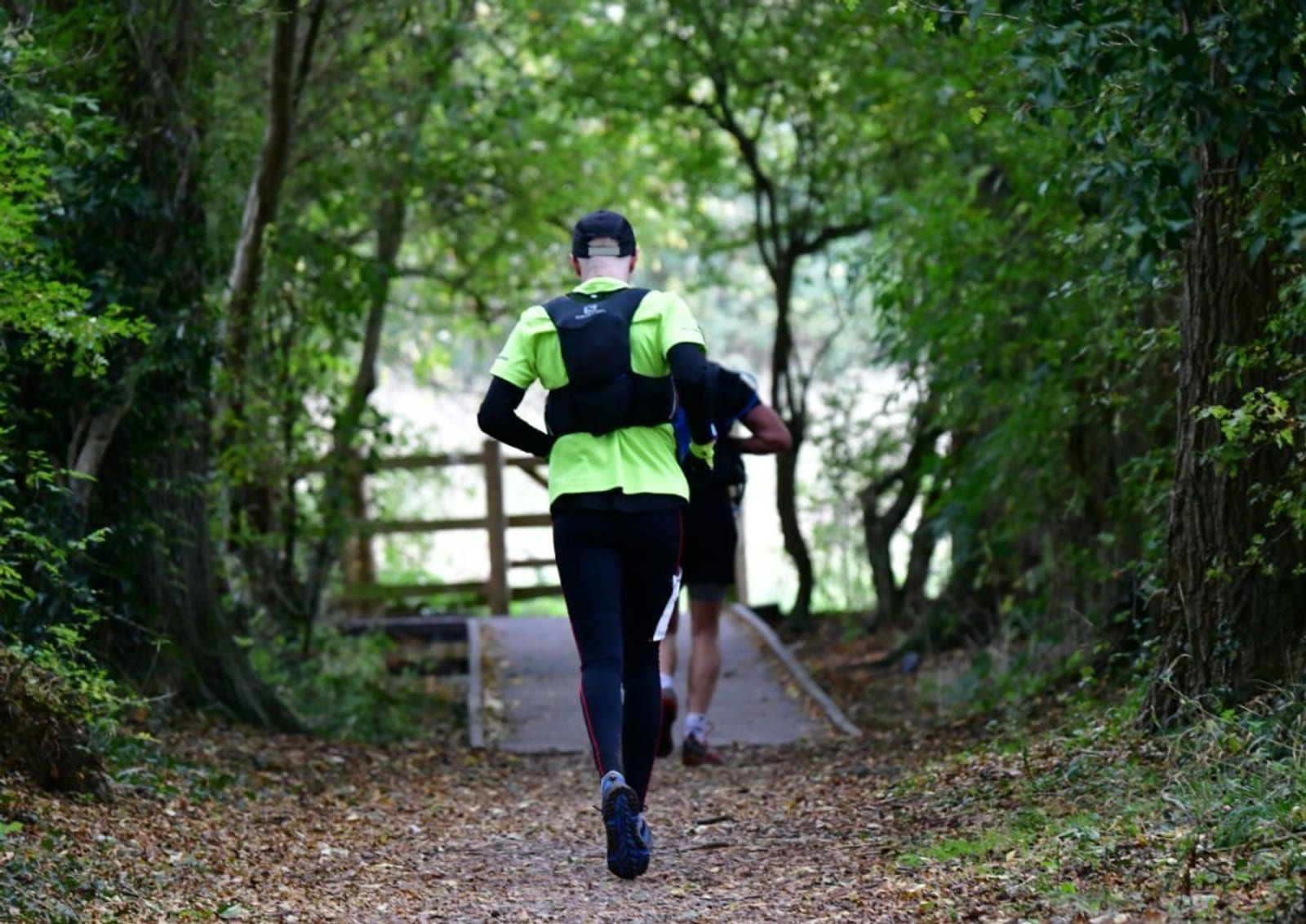Two joggers run down a wooded trail. The trail is surrounded by dense greenery and trees, with leaves scattered on the ground. One runner wears a bright neon green top and a black cap, while the other is dressed in darker attire, partially obscured by the first.