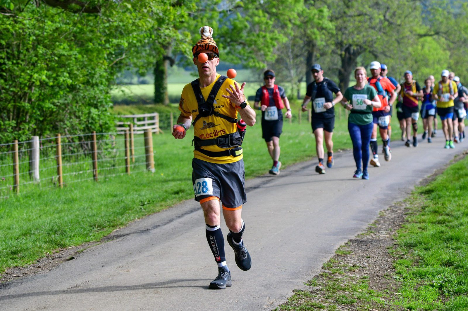 A runner in a yellow shirt and black shorts juggles three balls while running on a paved path during a race. He wears a quirky hat and has a race bib with the number 28. Other runners are behind him on a tree-lined path. It is a sunny day with vibrant greenery.