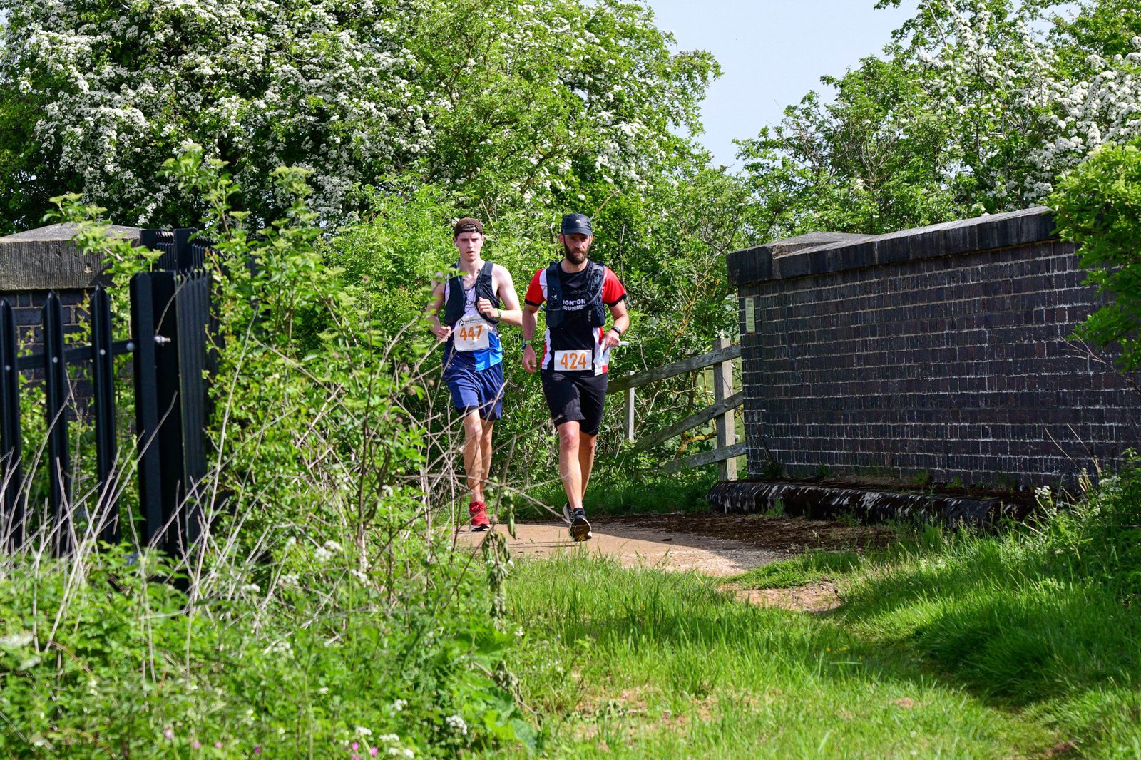 Two runners are participating in a race, running on a path surrounded by lush greenery and flowering bushes. They are wearing race bibs, athletic gear, and caps. The weather appears sunny, and a brick structure is visible to the right of the path.