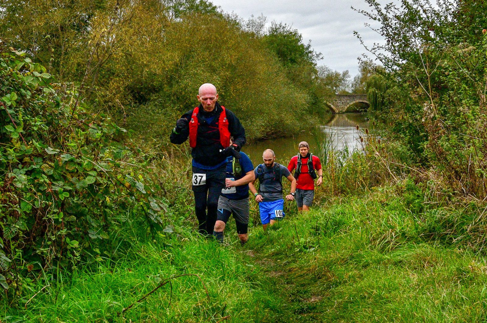 Four people are trail running on a grassy, narrow path alongside a river surrounded by dense greenery. One runner, closest to the camera, wears a red vest and has the number 37 on his shorts. A stone bridge is visible in the background under a cloudy sky.