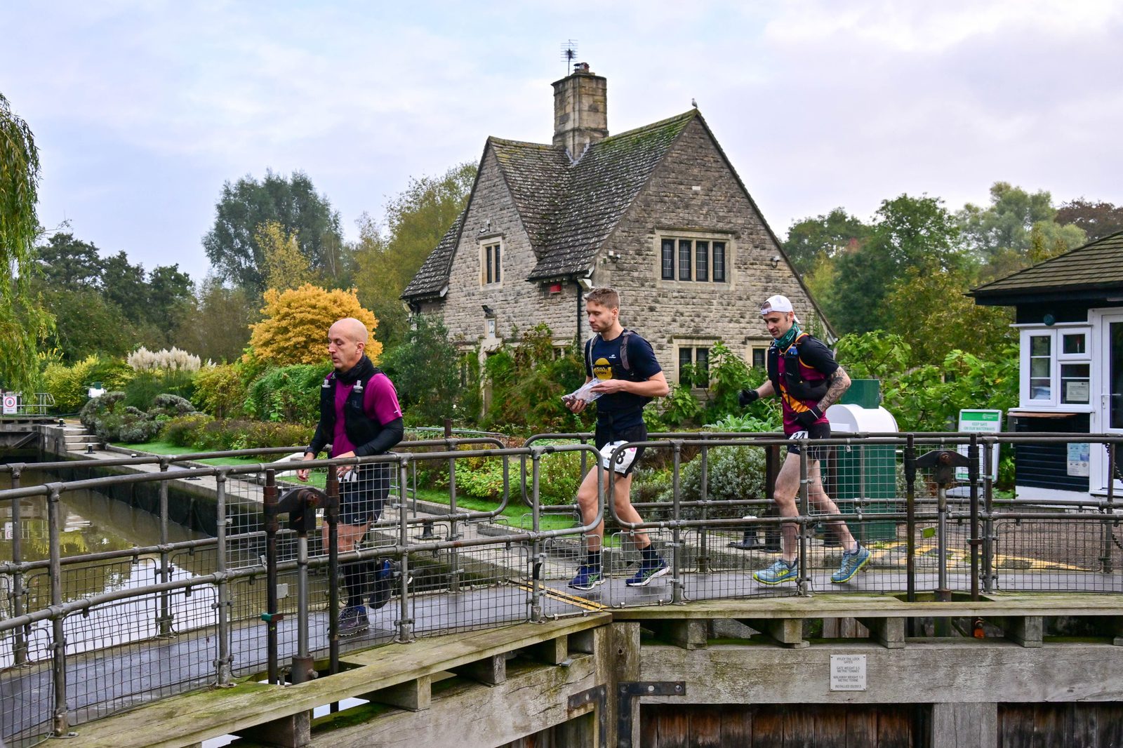 Three runners cross a metal bridge in front of a stone cottage with a chimney, surrounded by greenery. The runners are wearing race bibs and athletic gear. The scene is tranquil, with trees and a river visible in the background.