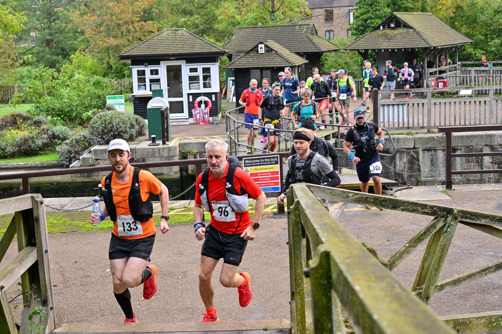 Runners in an outdoor race cross a wooden bridge. Two men in red shirts lead the group, wearing numbers 133 and 96. Behind them, other participants and spectators are visible near a building with a white picket fence. Trees and greenery surround the area.