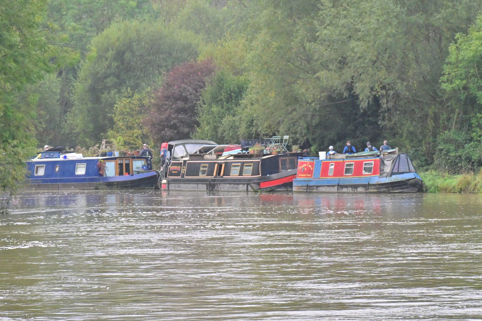 Boats moored side by side along the edge of a calm river, with lush green trees and shrubs in the background. People are visible on some of the boats, and the scene appears peaceful and tranquil.