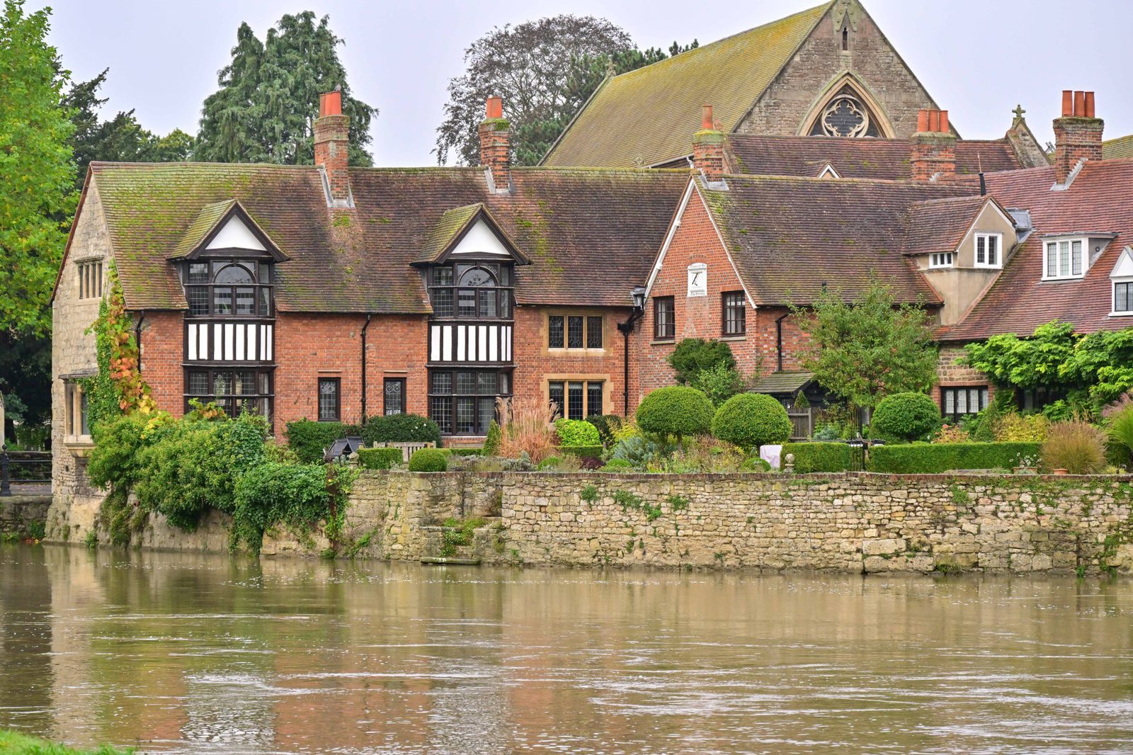 A serene riverside scene featuring red brick and Tudor-style houses with gabled roofs and chimneys. The houses are bordered by a stone wall and greenery along the water's edge, with trees and an old church visible in the background.