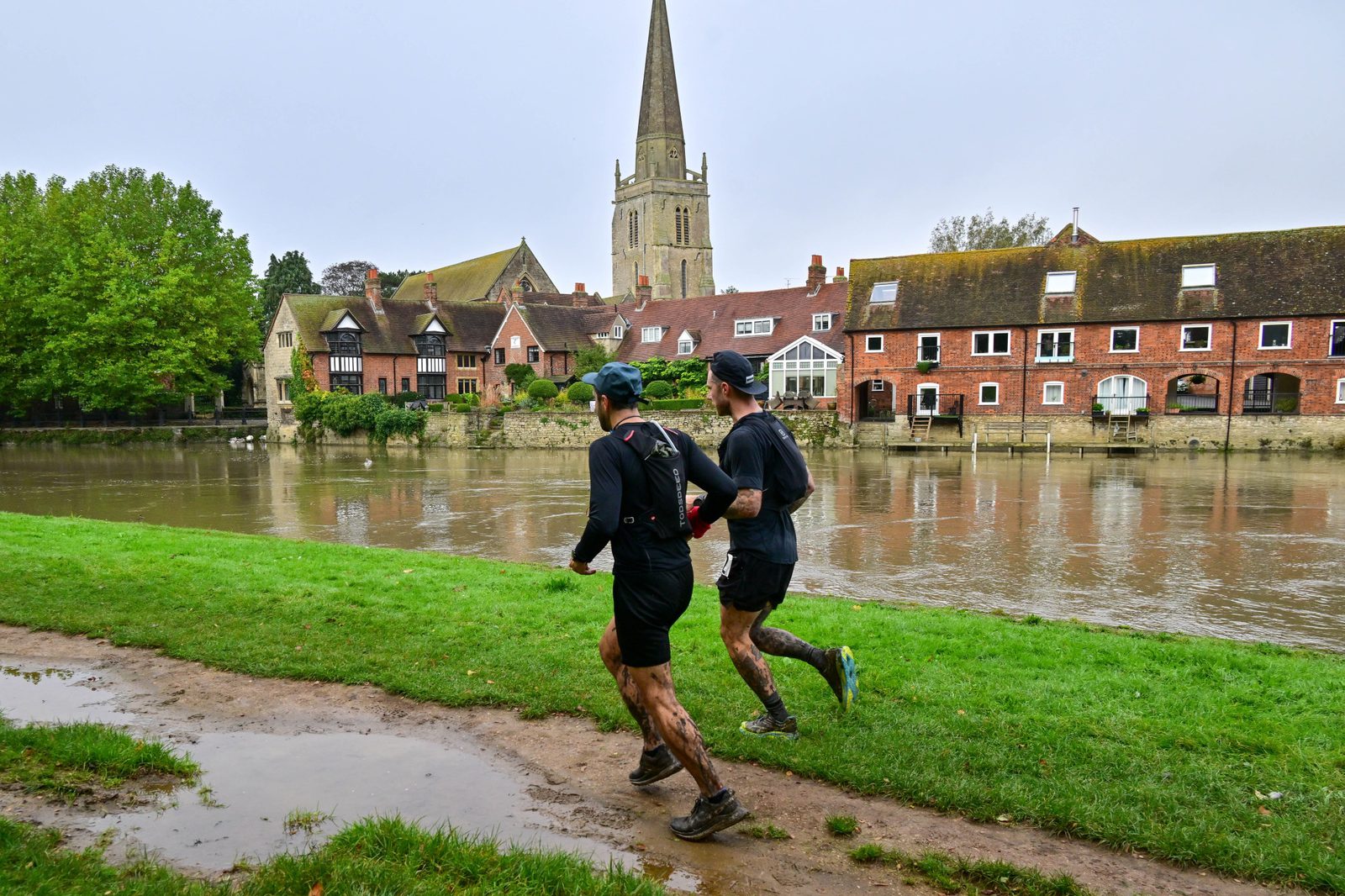 Two runners in black athletic wear and hats jog along a muddy riverside path with a church spire and old buildings in the background. The ground is wet, and their legs show signs of mud splashes. The river flows calmly beside them.