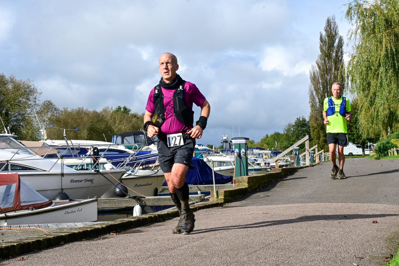Two men running beside a marina with various boats docked. The front runner, wearing a pink shirt and numbered 127, appears focused while the man behind him in a bright yellow-green shirt follows at a distance. Trees and a blue sky are in the background.