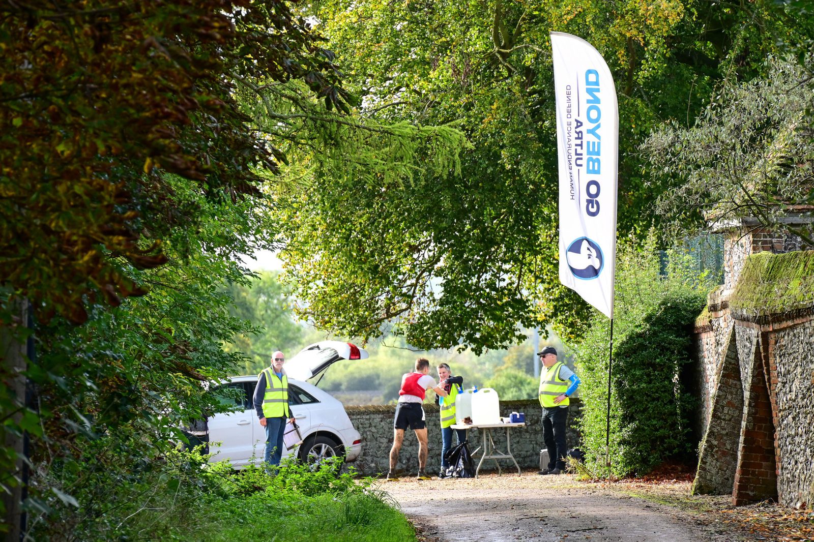 Three volunteers in yellow vests standing by a hydration station along a path near a white vehicle with its trunk open. One volunteer is assisting a runner in a red top and black shorts. A tall flag with “Go Beyond” branding is visible on the right. Trees line the path.