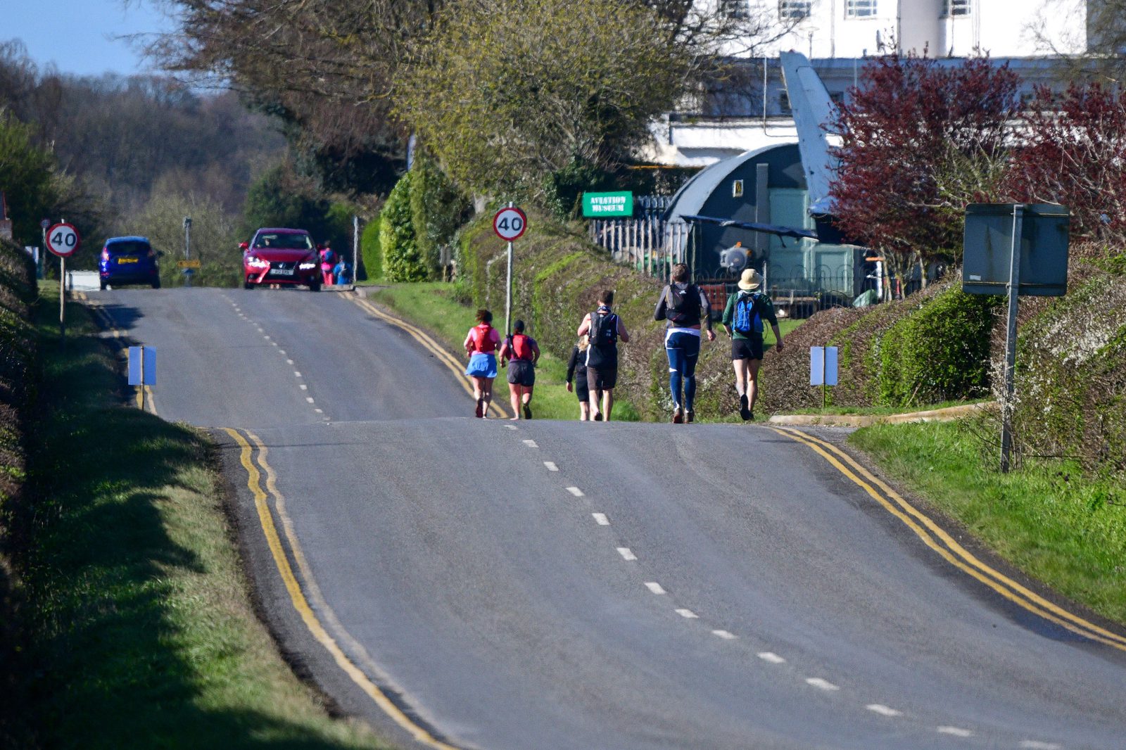 A person on a bicycle and four walkers are on a sunny roadway, heading towards a building surrounded by greenery. A red car is parked on the left side of the road, and there's a 40 mph speed limit sign visible. The road has double yellow lines on both sides.