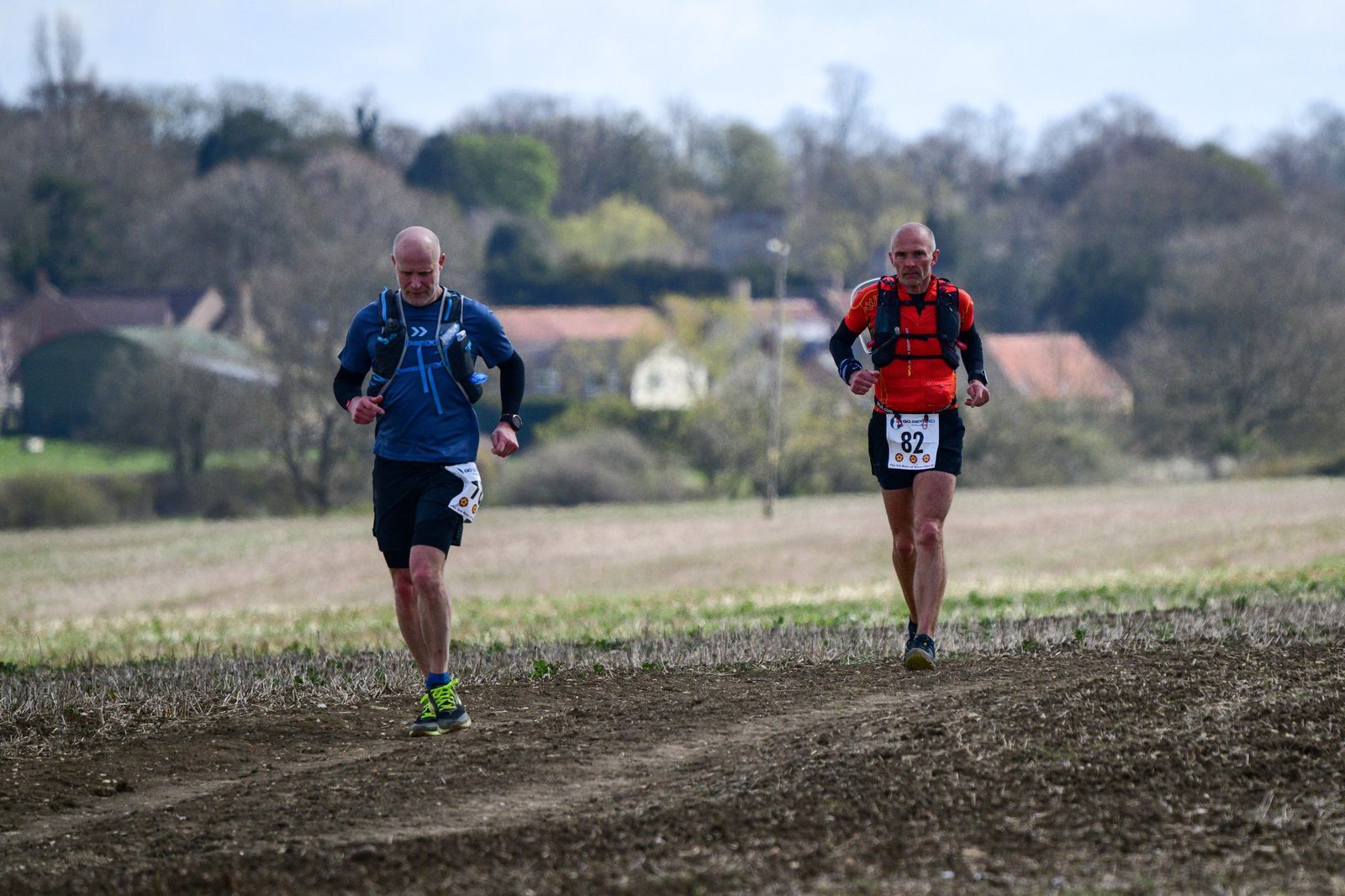 Two runners are participating in an outdoor race on a dirt path through a rural area. They wear numbered bibs, with the runner on the right wearing bib number 82. The landscape features fields, trees, and distant houses under a cloudy sky.