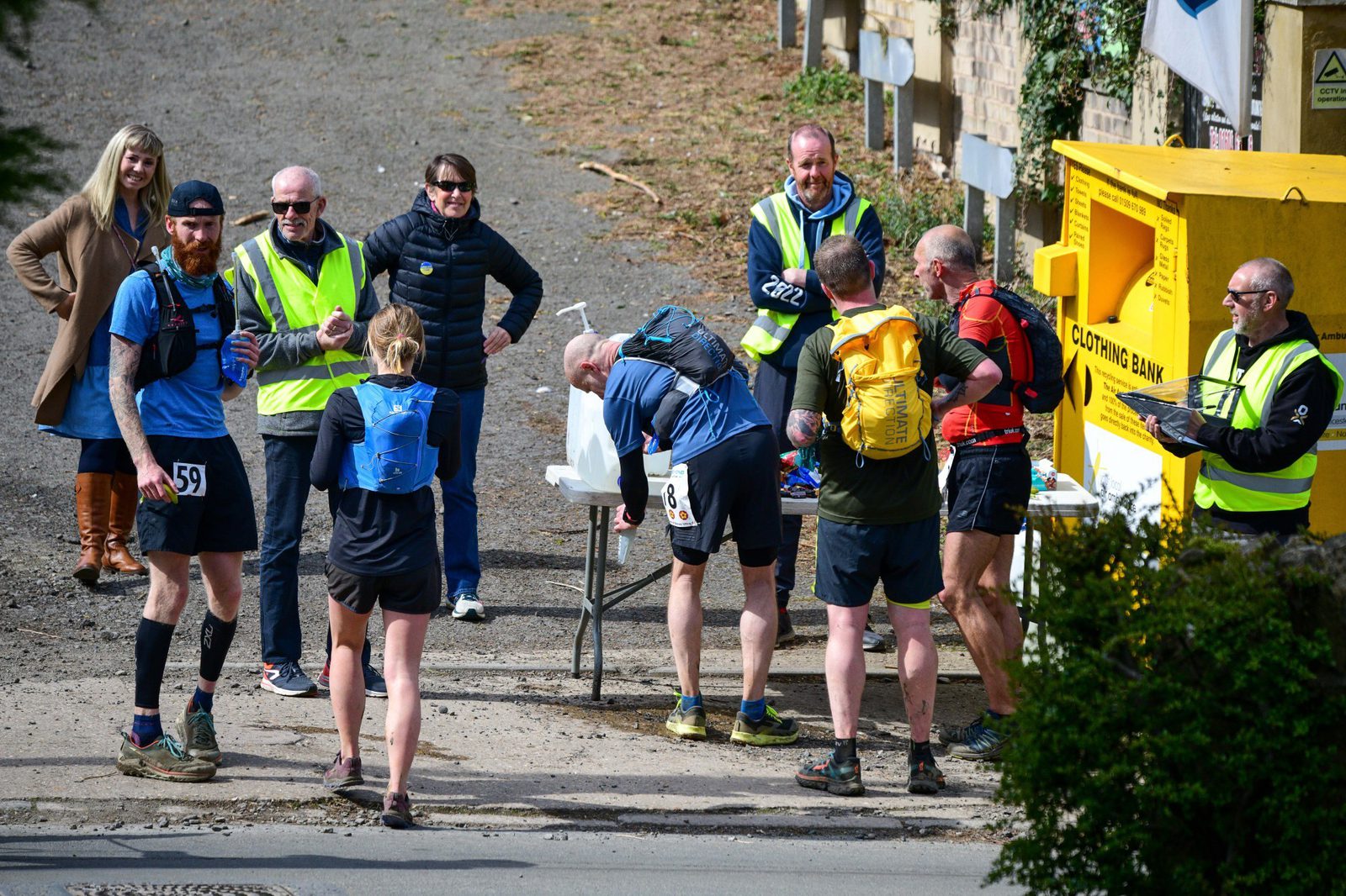 A group of runners is gathered at a hydration station during a race. Some are refilling bottles, while others are interacting with volunteers and spectators. The runners wear blue, yellow, and black athletic gear. A yellow clothing bank and a building are visible in the background.