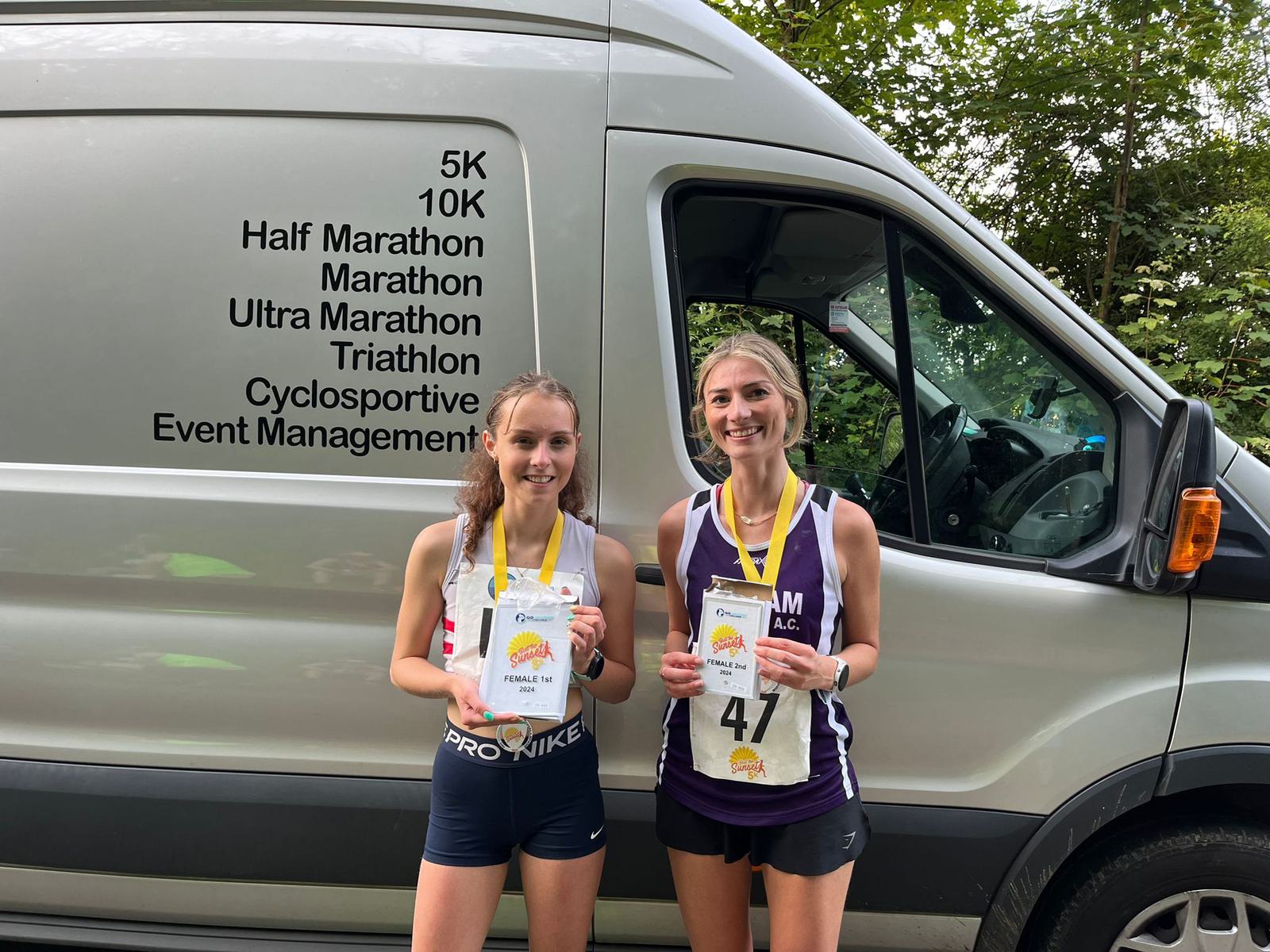 Two smiling runners with medals stand in front of a van. The van displays text listing various race events like 5K, 10K, and marathon. The runners each hold a certificate and wear athletic attire.
