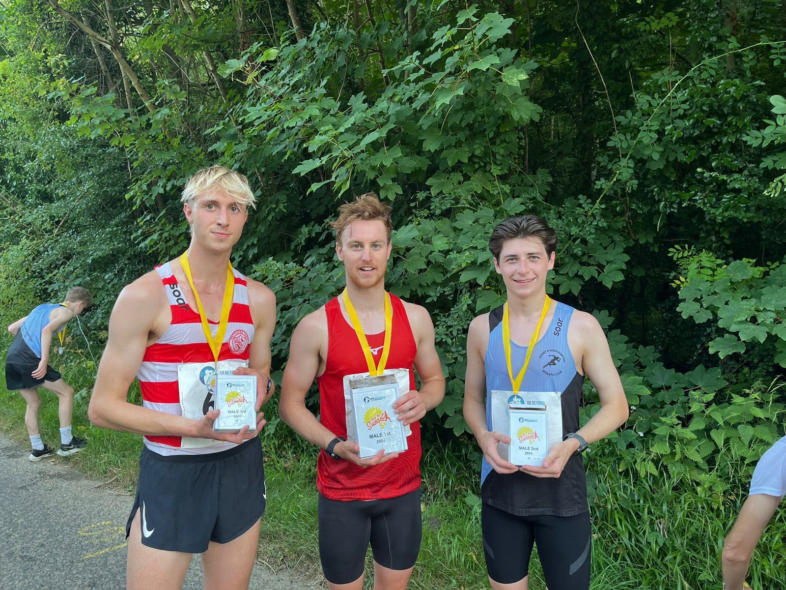Three runners wearing medals stand side by side holding plaques in front of a backdrop of lush green trees. They are dressed in athletic gear, each in a different color, and appear pleased with their achievements.