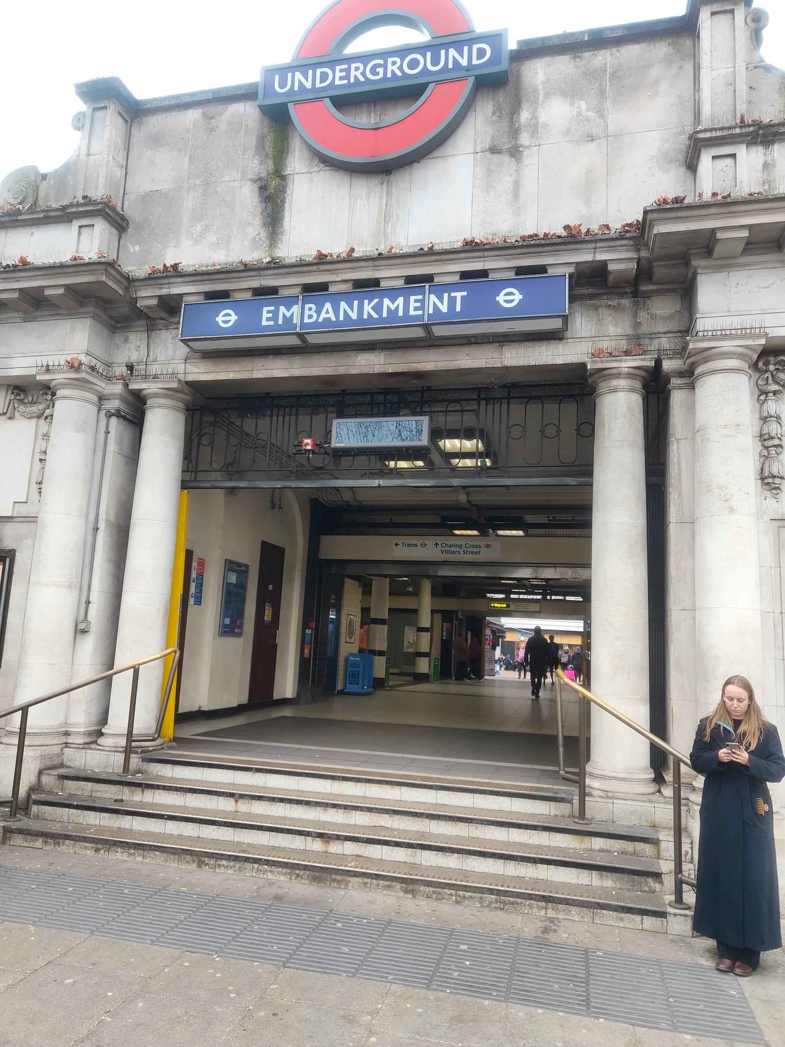 Entrance to Embankment Underground station with signage and stone columns. A person stands to the right, looking at their phone. Two ramps lead into the station where several people are visible in the background.