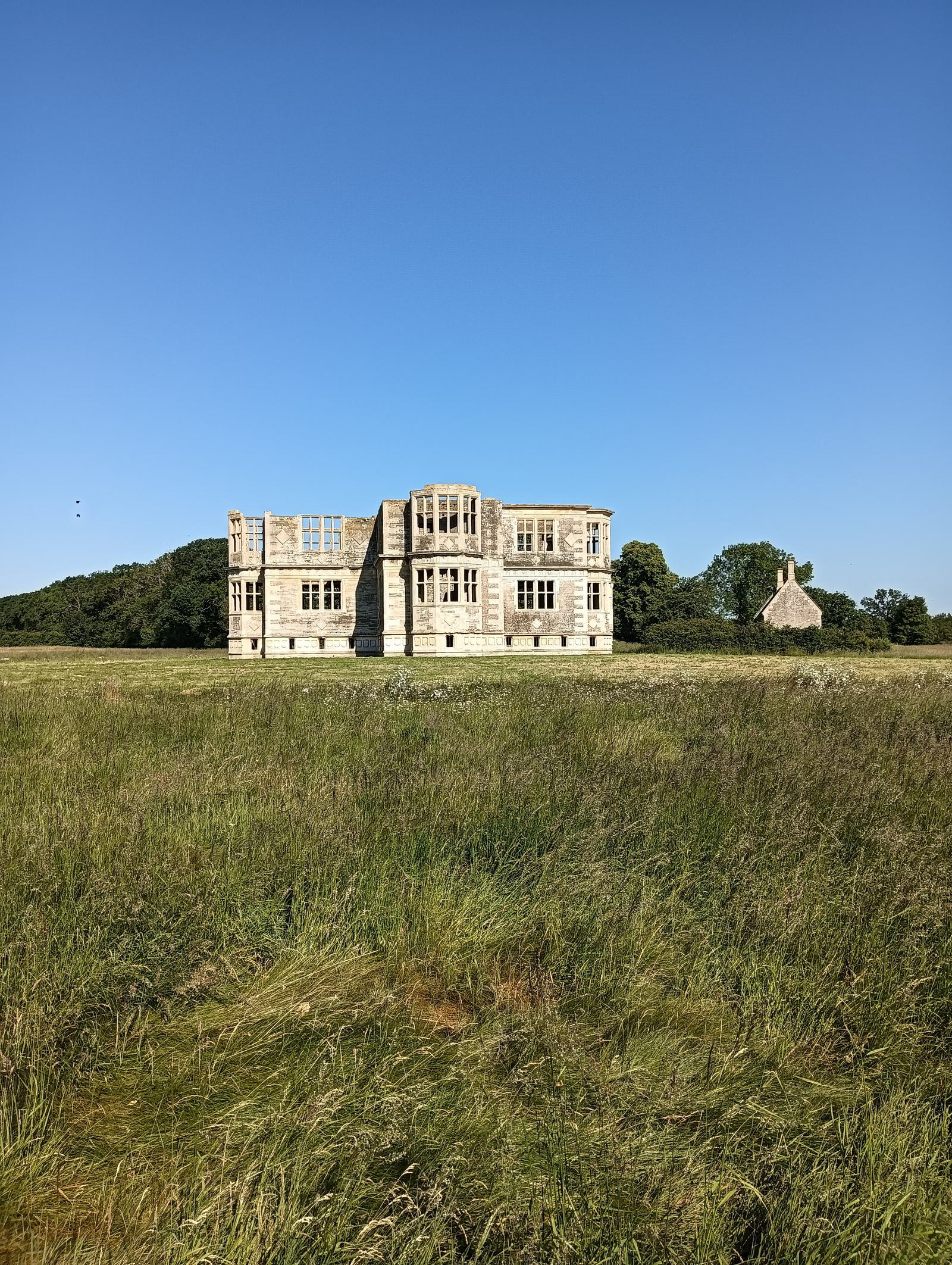 A solitary, partially ruined stone mansion stands in the middle of a vast grassy field under a clear blue sky. A small cottage is visible in the distance to the right, with trees and a hill in the background.