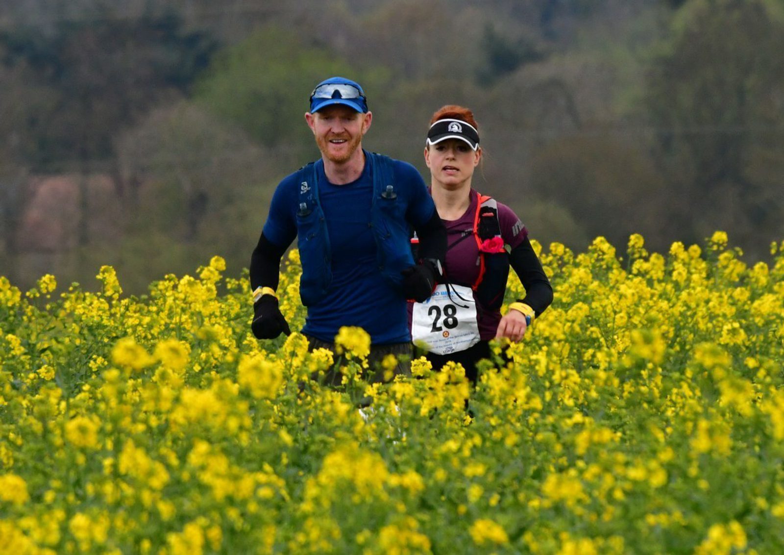 Two runners in athletic gear jog through a field of yellow flowering plants. Both wear race bibs—one with the number 28. The background is blurred but appears to be a mix of trees and distant terrain, suggesting a rural setting.