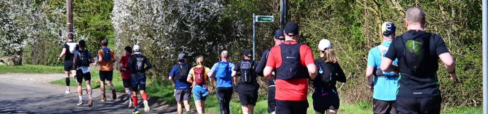 A group of people wearing athletic clothing and backpacks are running outdoors on a sunny day, surrounded by green trees and blooming bushes along a road or trail.