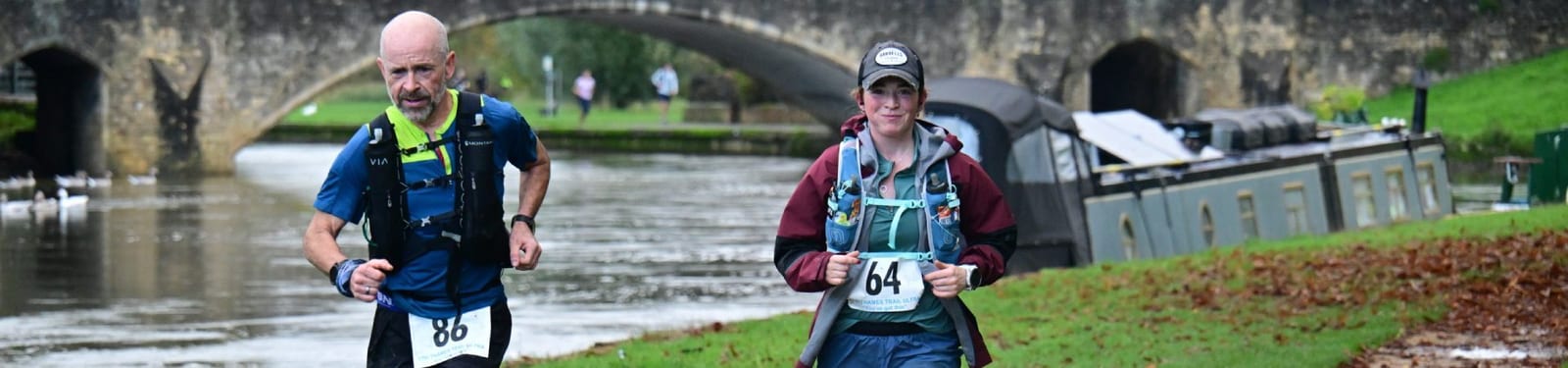 Two runners with race bibs jog along a canal path lined with fallen leaves, with a stone bridge and a narrowboat in the background. The runners appear focused, dressed in outdoor running gear.