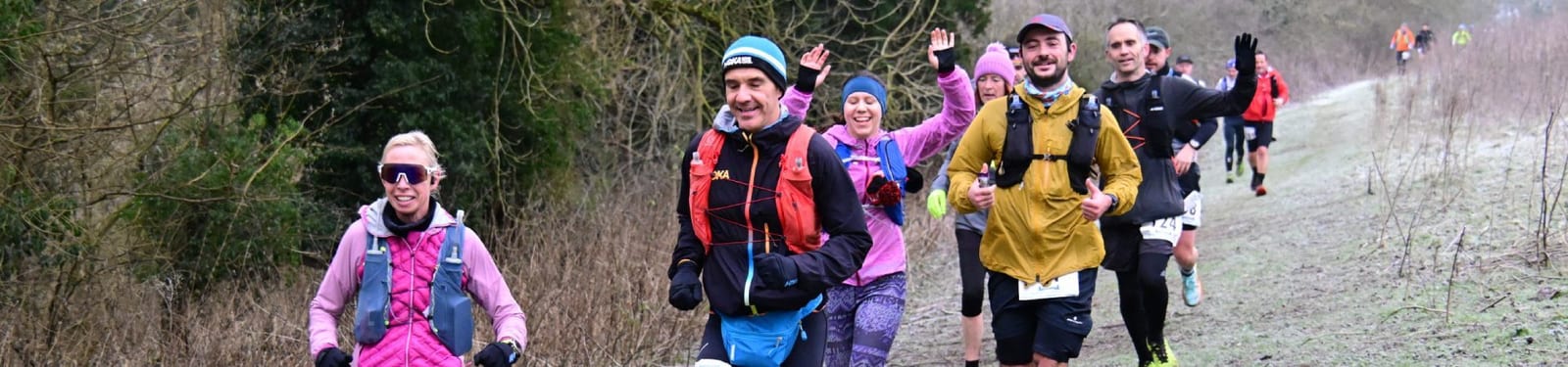 A group of runners in colorful winter gear jog along a grassy, frosty trail bordered by trees. Some runners smile and wave, enjoying the outdoor race on a chilly day.