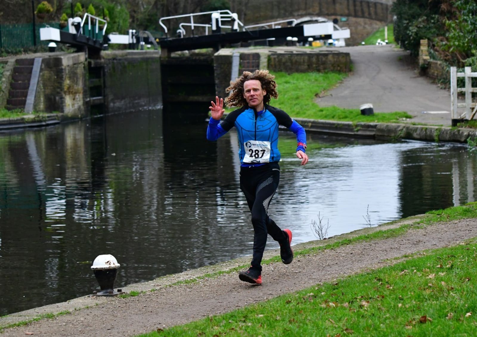 A runner wearing a blue jacket and race bib number 287 jogs on a path beside a canal, waving with one hand. A canal lock and green grass are visible in the background.