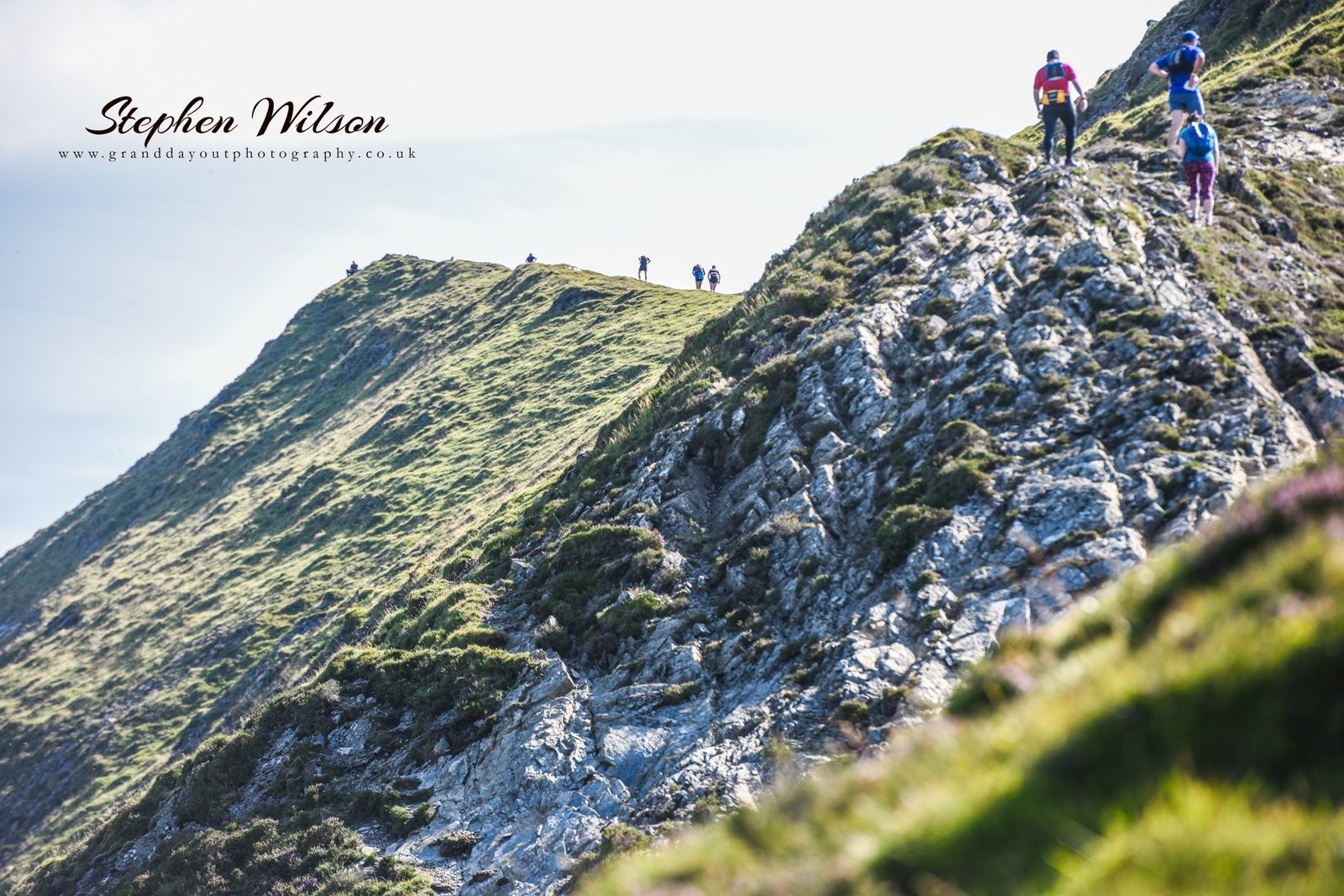 A group of hikers ascends a steep, rocky green hill under a clear sky. The rugged terrain is a mix of grass and jagged rocks. The image conveys a sense of adventure and natural beauty. The photographer's watermark is present in the top left corner.