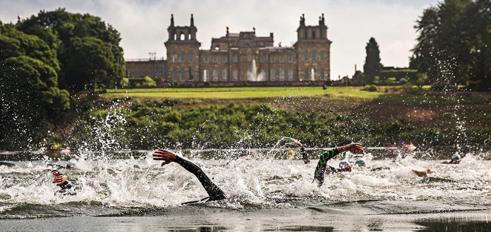 Swimmers in wetsuits compete in a race in a lake, creating splashes as they move. A grand, historic building stands in the background amidst lush greenery, with a clear sky above.