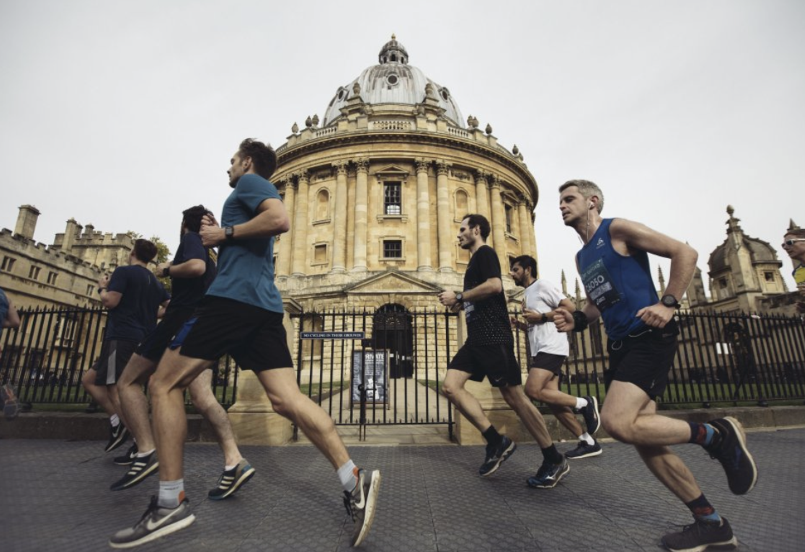 A group of runners jogs in front of the Radcliffe Camera, a historic building in Oxford, England. The runners wear athletic attire and appear to be participating in a race or organized run. The sky is overcast, and the architectural details of the building are prominent.