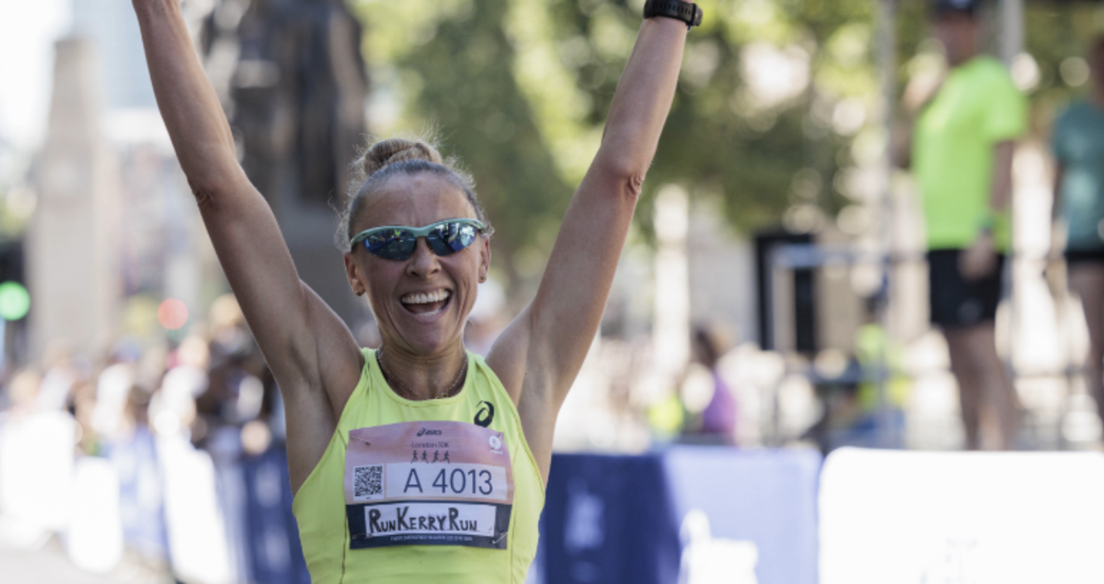 A person wearing sunglasses and a bright yellow tank top with a race bib "A 4013" and "RunKerryRun" is raising their arms in triumph. The background shows crowds and other participants in a city marathon setting.
