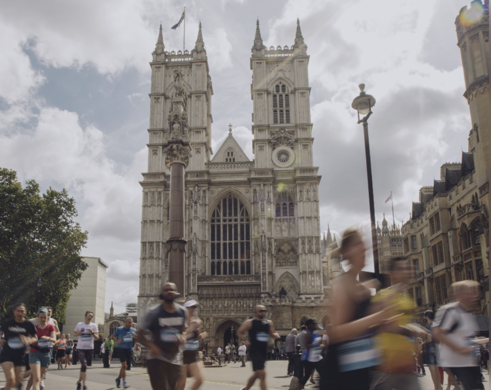 Runners pass by a large Gothic-style cathedral with two prominent towers in the background. The sky above is partly cloudy. Trees, buildings, and lamp posts are also visible in the scene.