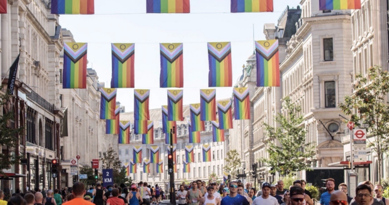 A bustling street scene during a parade or event in a city. Colorful rainbow flags hang overhead, spanning the street between buildings. The crowd below comprises people walking and engaging in various activities. Trees line the street, adding a touch of greenery.