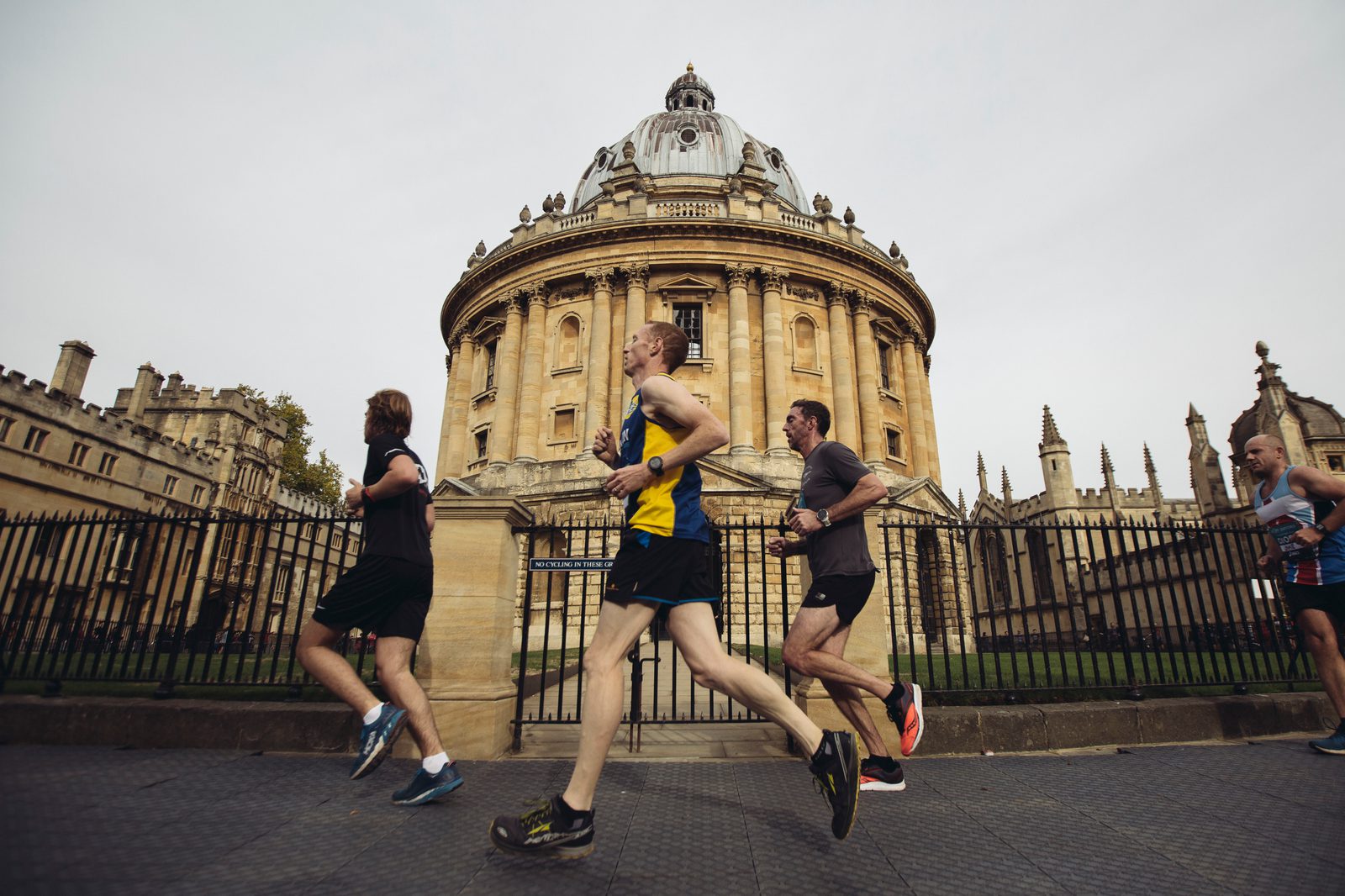 Four runners jog past the Radcliffe Camera, an iconic circular library building with a dome in Oxford, UK. The runners are dressed in athletic wear, and the historic architecture provides a striking backdrop to their energetic activity.