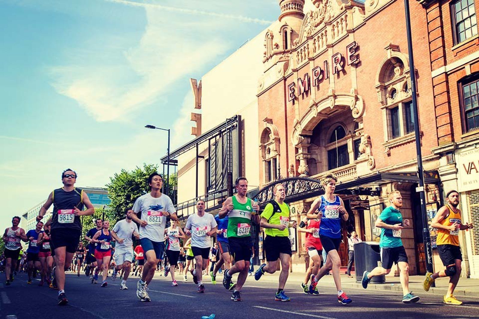 A diverse group of runners participates in a marathon on a sunny day. They are passing a historic red-brick building with the word "EMPIRE" displayed prominently at the top. The runners wear various colorful athletic outfits and race bibs.