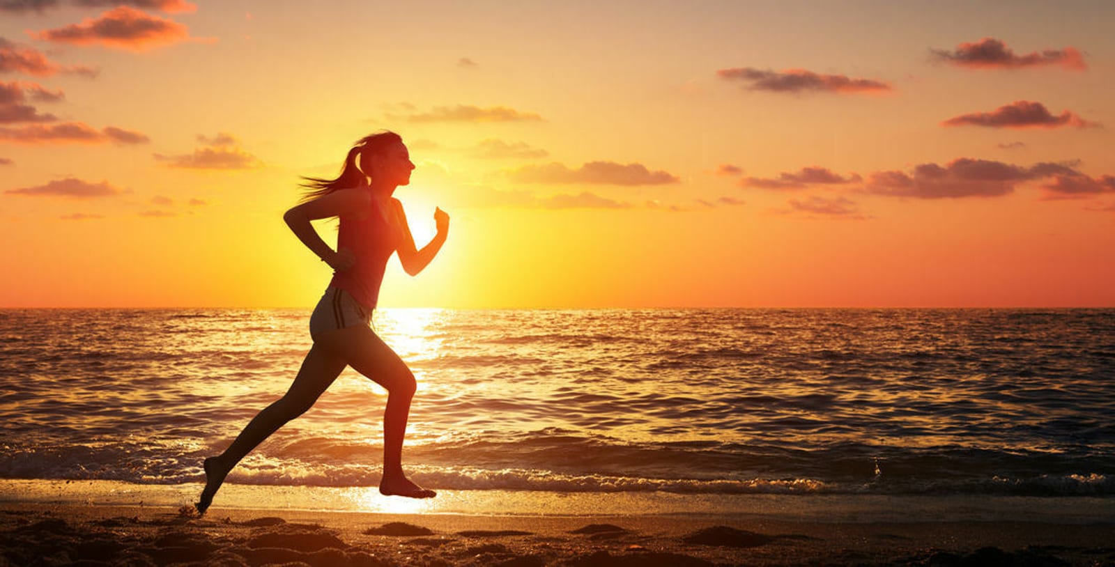 A woman is running along a beach during sunset. The silhouette is against a backdrop of an orange and yellow sky, with the sun low on the horizon and a calm ocean reflecting the sunlight. She has her hair in a ponytail and is wearing a tank top and shorts.