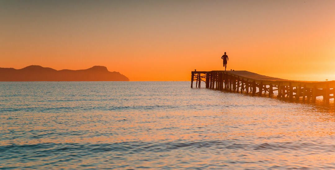 A person stands at the end of a wooden pier overlooking a calm sea during sunset. The sky is painted in warm orange and yellow hues, and distant, silhouetted mountains create a serene backdrop. The water gently ripples towards the shore.